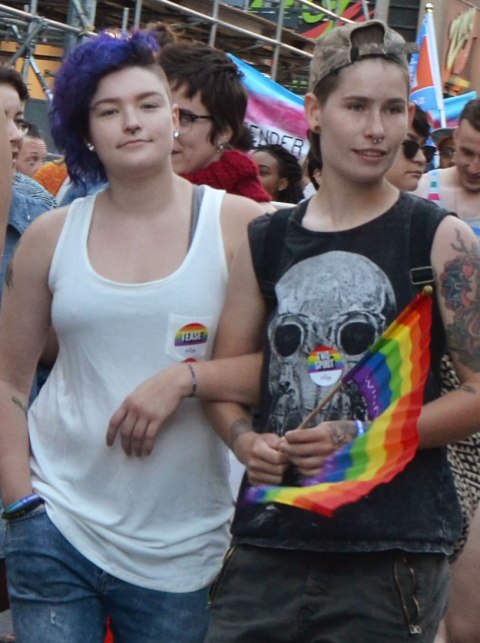 a couple walking arm in arm in a parade, one has short blue hair and the other is wearing a t shirt with a skull on it and holding a rainbow pride flag