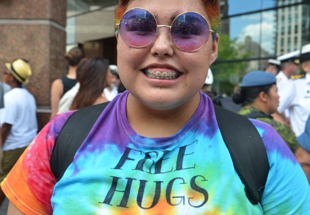 A young person with very short hair, wearing round purple and pink sunglasses and a tie dyed T-shirt that says Free Hugs on it. 