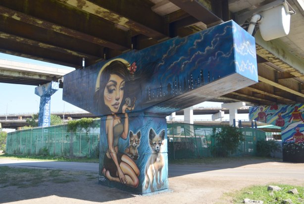 A mural on a bent in an underpass, a young woman is kneeling. She is holding a mouse in one hand. Two foxes stand beside her. 