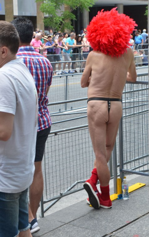 A man in a large red afro wig is standing beside the parade route, on the sidewalk but behind the barricade. Viewed from the back, all he is wearing is a leather thong and red shoes and socks.