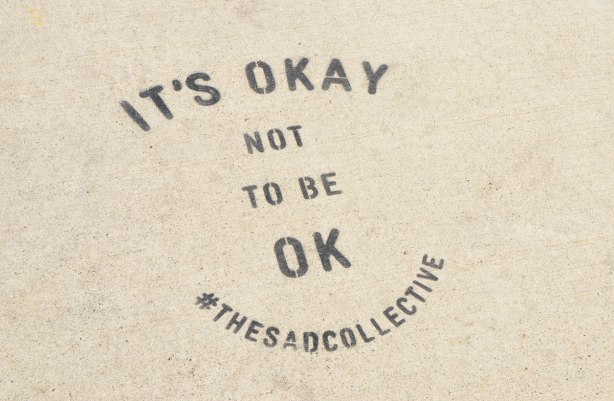 words stenciled onto the sidewalk in black, that say It's Okay not to be OK #thesadcollective