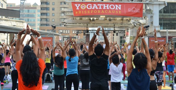 A banner that says Yogathon rise for a cause hangs over a crowd who are standing with their arms in the air as part of the activity