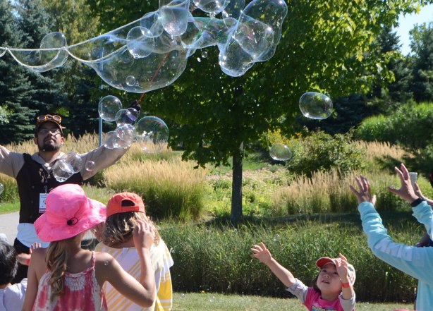 very large bubbles being made in front of a crowd of children and adults, kids chasing and trying to catch and burst the bubbles, small girl with her arms outstretched 