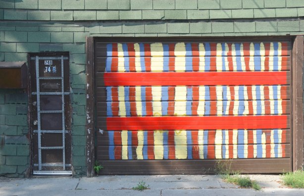 a garage door painted in yellow, red and blue vertical stripes as well as two wide horizontal stripes, the walls are green shingle and the entry door is black with a grid of metal stripping on it. 