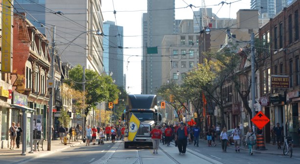 looking east on Queen Street West towards the Labour Day parade. A large truck is in the middle of the road, and people waving flags are walking in front of it. Lots of people walking beside it and behind it. Stores on Queen Street to the sides, taller buildings in the background. 