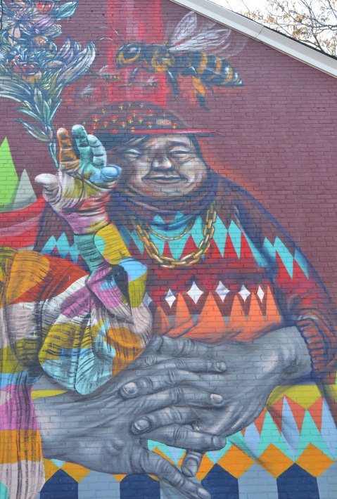 part of a larger mural of elicser and Troy Lovegates, colourful people, a heavy set man with a cap on, seated with large hands folded on his lap, wearing a sweater with rows of colourful diamond shapes. A large bee is flying above his head. 