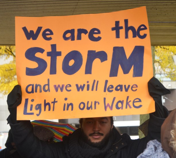 people at a rally protesting against Donald Trump as President of the USA, a man holds up an orange sign with black letters that say We are the storm and we will leave light in our wake 