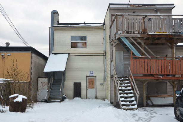 the back of two buildings, one has undeisturbed snow behind it and a sign over the door that says R Coin Laundry. The building beside it (and attached to it) is three storeys high. The upper two levels have a wood railing and balcony out the back. Snow on the ground but lots of foot steps in the snow. 