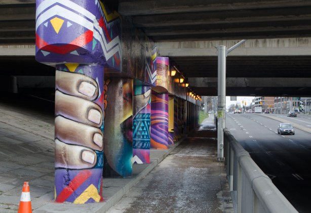 pillars and supports under an overpass that have been painted in bright colours by smoky and shalak - a large face in the center pillar, with hands gripping the outer pillars on each side of the face