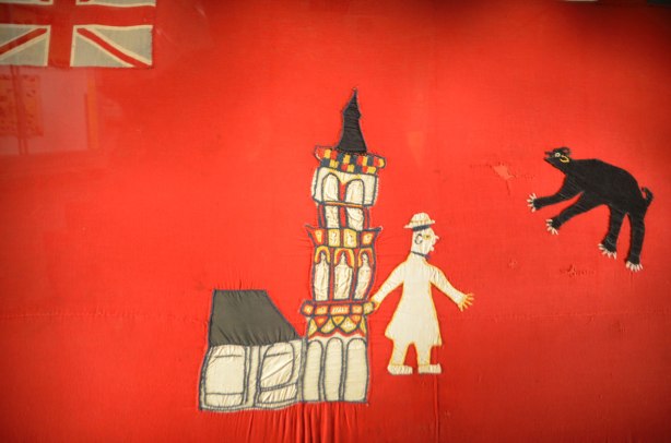 close up of a flag, hand made, British Jack in the top left corner, a man walks in front of a church in the center, a black bear in the top right. 