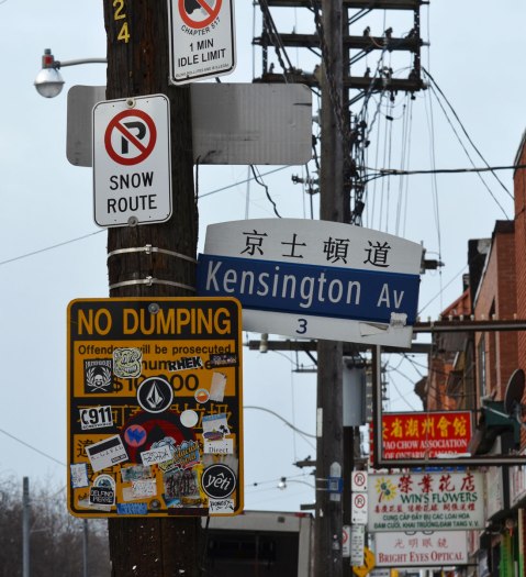 two hysro poles, one with a lot of signs on it and the other with three horizontal wood sections at the top of it. Signs are a street sign for Kensington Ave in both English and Chinese, a yellow no dumping sign that has been covered in stickers, and a no parking sign. Signs for Chinese businesses are in the background. 