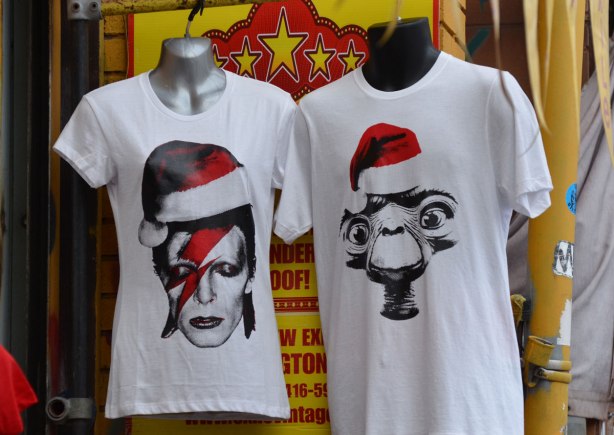 two t-shirts on display outside a store, both are white and both have heads of famous people wearing Santa hats. One is David Bowie and the other is E.T. 