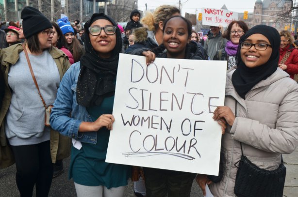 Three black women, one with a head scarf on, hold a sign that says Don't silence women of colour, part of a crowd at Womens March, toronto 