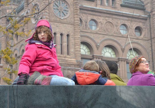 a young girl in pink sits on top of the War Memorial at QUeens Park, the parliament buildings are behind her. 