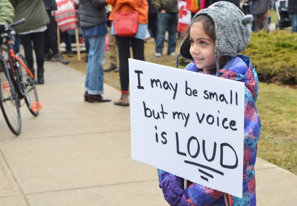 Womens March, Toronto, a young girl in a grey hat holds a white sign that says I may be small but my voice is loud. 