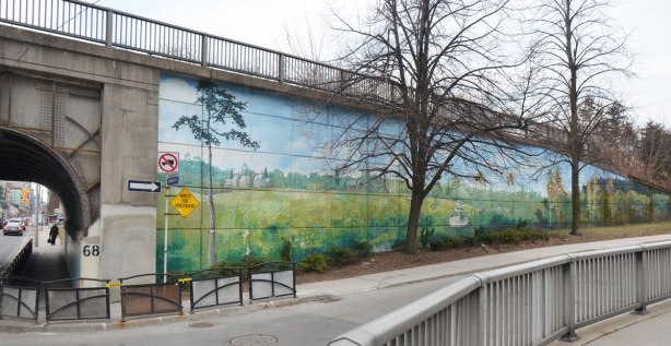 mural painted along the side of a wall that is part of the embankment for a railway bridge Mural is a country scene, grass and fields, a farm in the distance and a couple of trees. 
