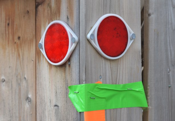 two round red reflectors mounted on a wood fence, look like two eyes, a piece of green tape is also on the fence in such a way that it looks like the mouth