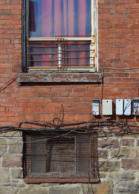 two windows on a red brick house with stone foundation, basement window and first storey window. The upper one has a red curtain