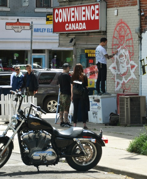 motorcycle in the foreground, a man painting a white outline of a rose as part of a mural on the side of Canada Convenience store on Queen West, a few people watching him paint 