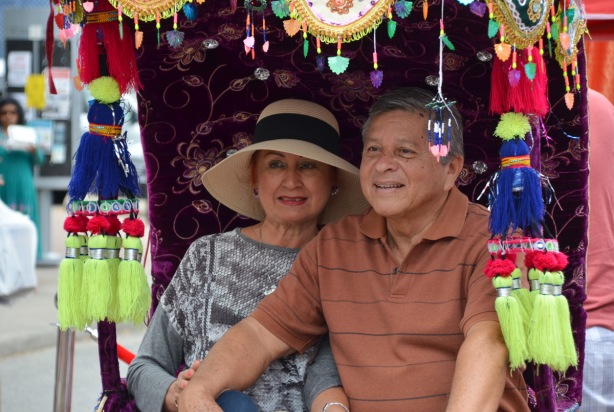 an older couple pose for a picture while they are sitting in a decorated cart, purple velvet, and many colourful tassles