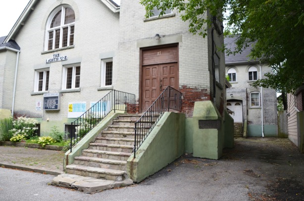 steps up to a large brown door, one of the entrances of the Toronto Gospel Lighthouse church, a greyish white building with another building behind. 