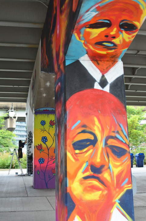 conrete pillar in Underpass park that has been painted with large orangish toned faces, by Carlos Delgado