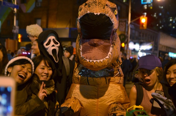 a large inflatable dinosaur costume, a death ghoul costume, and many people standing around them posing for photos at a night time halloween party on Church St., toronto