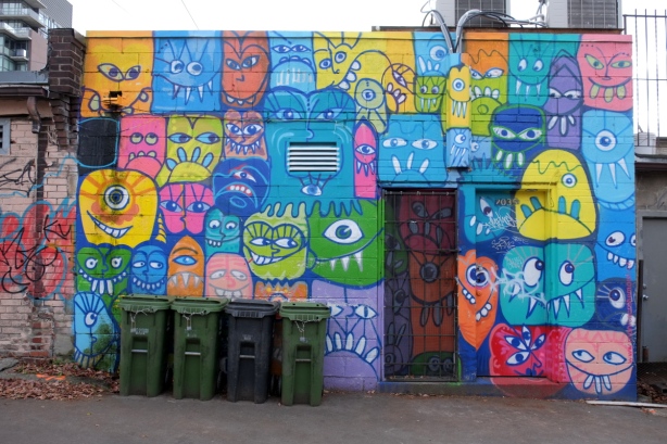 exterior wall in a laneway with mural by monicaonthemoon with many silly stylized faces in bright colours 