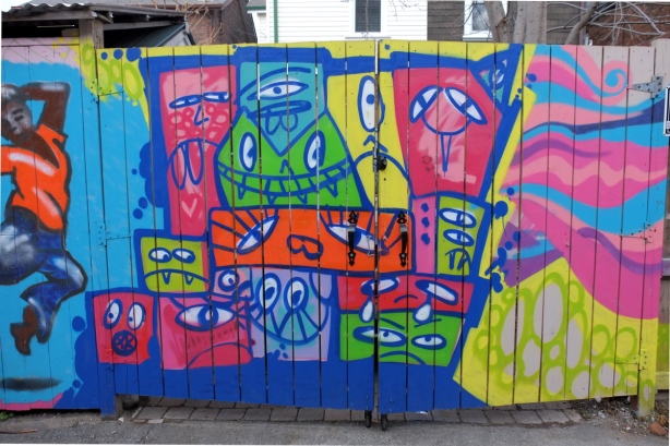 fence in a laneway with mural by monicaonthemoon with many silly stylized faces in bright colours 