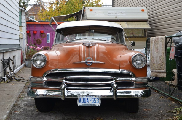 an old orangish brown Chrysler car parked in a driveway, front facing the street, 