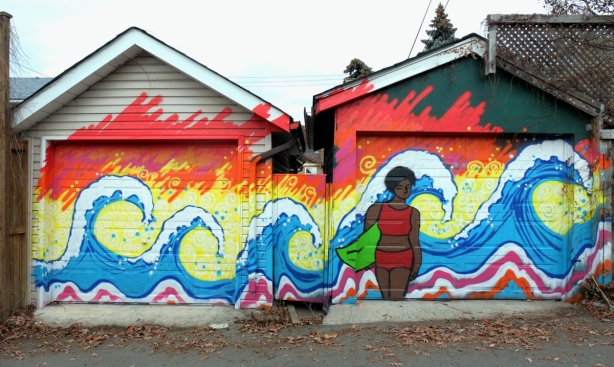 mural of a brown woman with a two piece bathing suit standing on a beach holding onto a surfboard. There are 4 very big waves behind her. Painted over two garage doors. 
