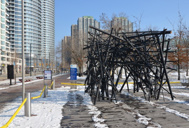 a tunnel like structure made of black bamboo poles loosely intertwined and joined together on the sidewalk beside Queens Quay, snow on the ground, condos in the background. 