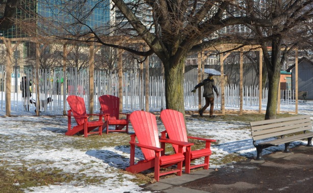 a man is touching and pushing a series of wind chimes that are part of an art installation at Toronto Music Garden as he walks by it. Snow covered ground, red muskoka chairs, park, large tree, 