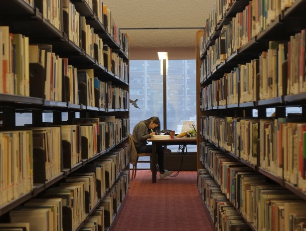 looking down an aisle between two stacks of books (book shelves), a woman is sitting at a table studying and writing, there is a window behind her 