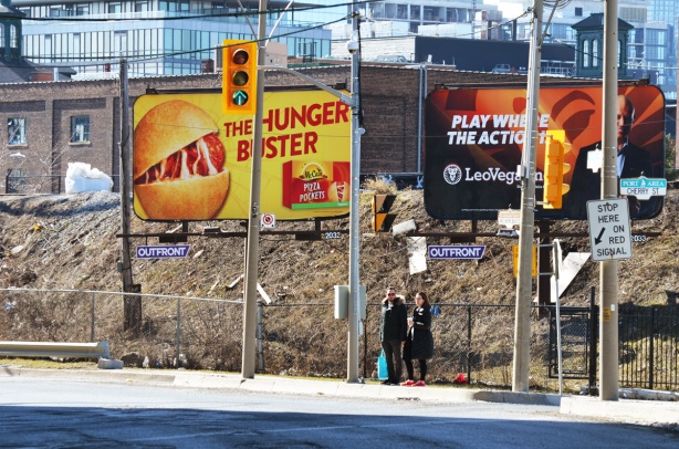 two people standing on a sidewalk as they try to figure out how to cross the street, two large billboards behind them, train tracks (elevated) behind that. 