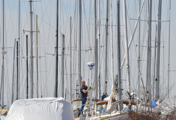 a lot of masts from sailboats standing upright, a man walks on one of the boats as he gets it ready to go back in the water after the winter