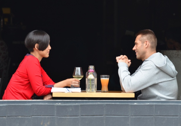 a couple sitting in a restaurant, across from each other, in the window, window is open, drinks on the table .  Woman has glass of white wine, man has something orange
