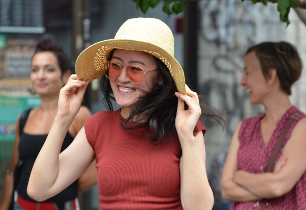 young Asian womanin a straw coloured wide brimmed hat, smiling as she dances, she is holding the brim of her hat with both hands so it doesn't fall off