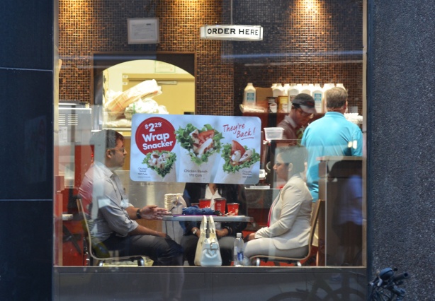 three people at Tim Hortons, sitting at a table. Seen from the outside, a poster on the window blocks the view of one of the people