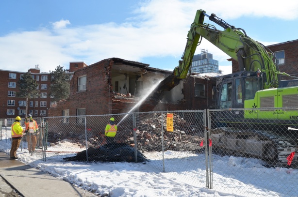 workmen spray water as a machine arm pulls apart a building that is in the process of being demolished