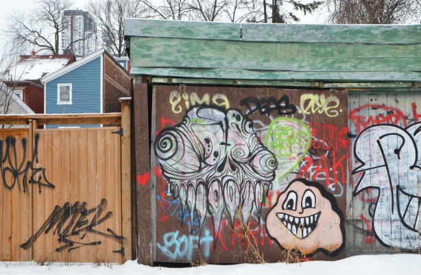 a grominator piece of graffiti and a pink smiley face monster, both on a brown garage door in an alley 
