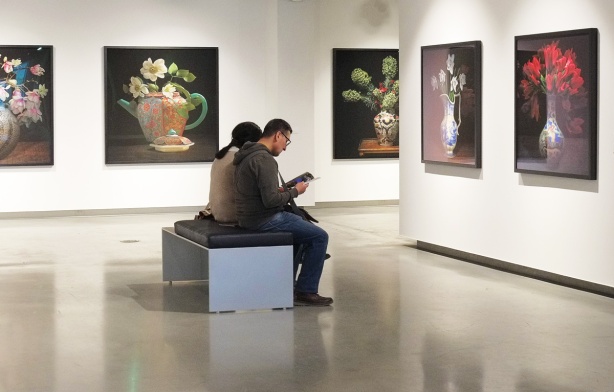 people sitting on a bench in a gallery, reading, large photos of flower bouquets on the walls around them 