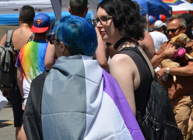 women at pride parade, one with blue hair with an asexual flag around her back - purple, white, grey, and black stripes 