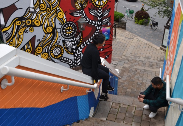 a young woman sits on the railing of an outdoor staircase that has been painted with street art. A man is taking a pictrure with his phone of her feet against the artwork