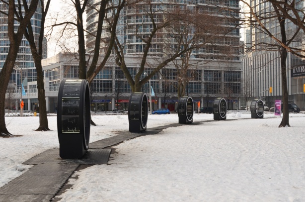 black circular structures that are part of an interactive art installation called Loop, arranged in a semi-circle at Yor Street Park, snow on the ground, trees with no leaves, no people there 