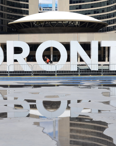 A couple stands behind the o in 3 D toronto sign, barriers in front of sign, most of the water has been removed from pool in front, so have puddles with reflections of sign and city hall 