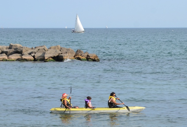 two women and a girl paddling in a yellow boat, a sailboat is in the background 
