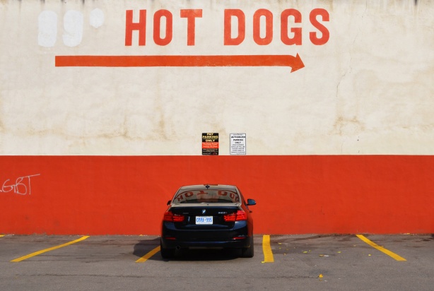 a black car is parked in a parking lot beside a wall that is orange on the bottom and beige on top. on the beige part is a large arrow pointing right with the words hot dogs 