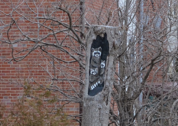 a tree trunk, about 6 feet high, with a section carved out and a raccoon family painted on it. 