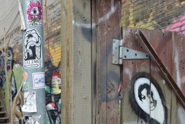 stickers on a pole, a daisy in pin, a feelings boi, beside a wood fence and gate with stencil graffiti of a man's face in a white oval framed in black 